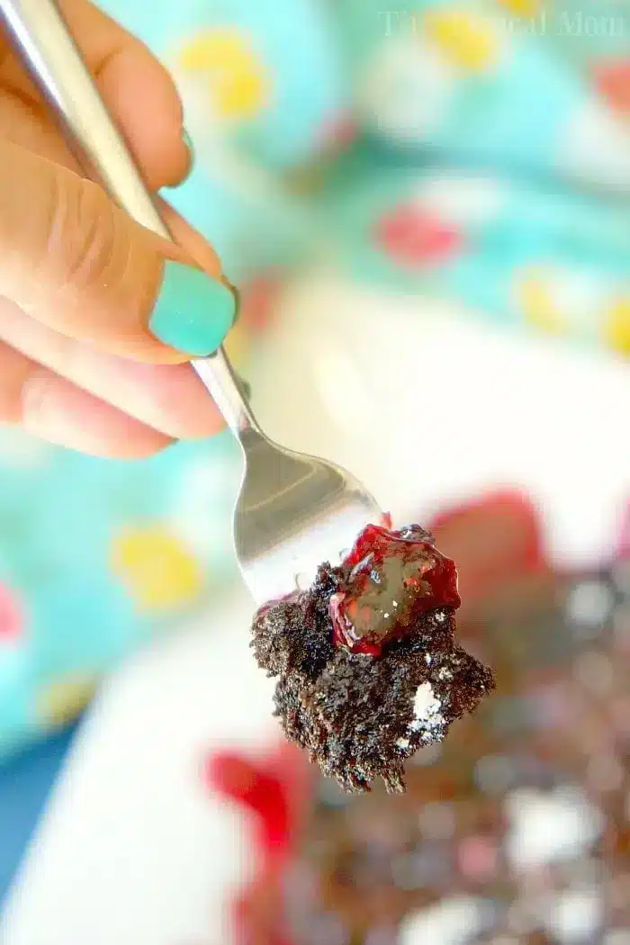 A hand with teal-painted nails holds a fork with a bite of instant pot chocolate cake topped with red berry sauce. In the background, a blurred colorful tablecloth is visible with more of the dessert on a plate.