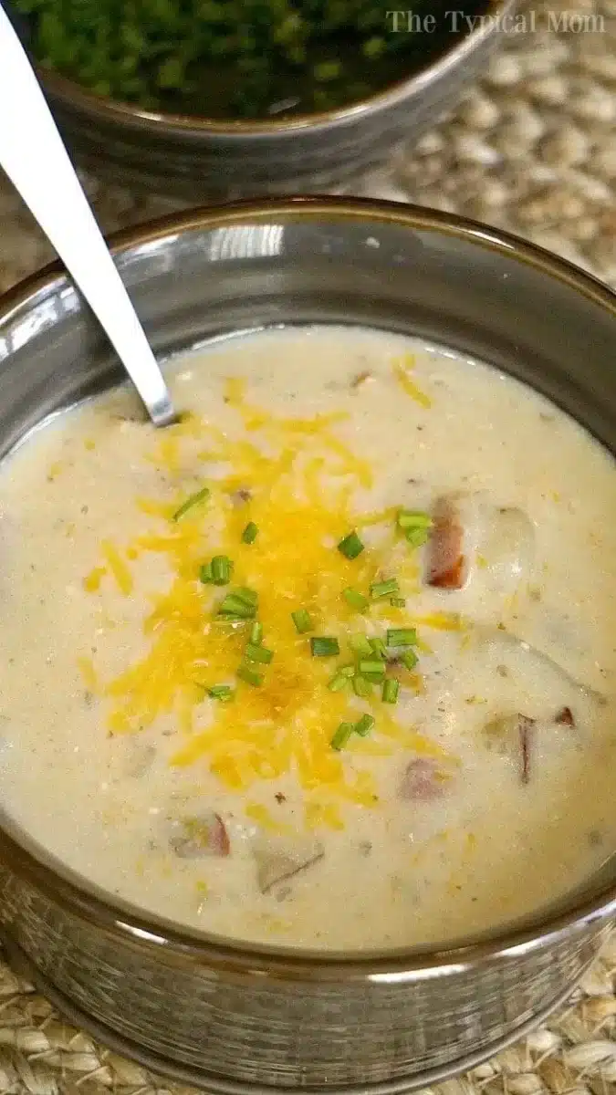 A bowl of creamy instant pot potato cheese soup garnished with shredded cheese and chopped chives. A spoon rests inside the bowl. The soup features small chunks of ingredients, possibly bacon or extra potato. The bowl is perched on a woven mat surface, inviting you to savor each comforting bite.