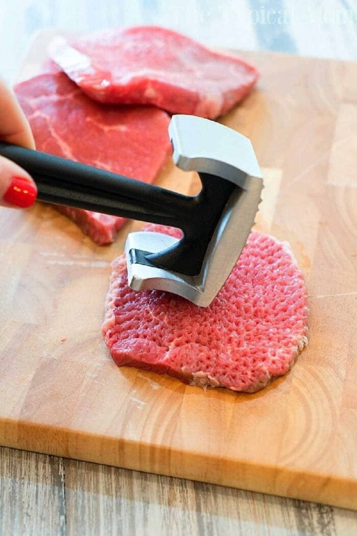 A person using a meat tenderizer mallet on a slice of raw beef destined to become cube steak, placed on a wooden cutting board. Two additional pieces of raw beef are in the background, ready for the slow cooker.