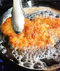 Metal tongs holding a golden, breaded cutlet sizzling in bubbling oil, reminiscent of the crispy goodness of an Air Fryer McDonalds Fried Chicken Sandwich.