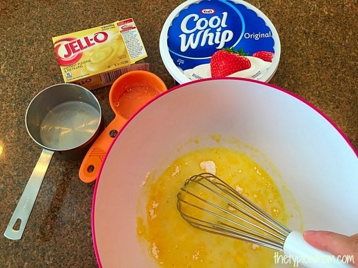 A mixing bowl containing a yellow mixture being whisked. Surrounding the bowl is a package of Jell-O vanilla pudding mix, a tub of Cool Whip, an empty measuring cup, an orange measuring spoon with a small amount of powder, and a metal tablespoon—essential ingredients that could elevate any dessert or chocolate mug cake recipe.