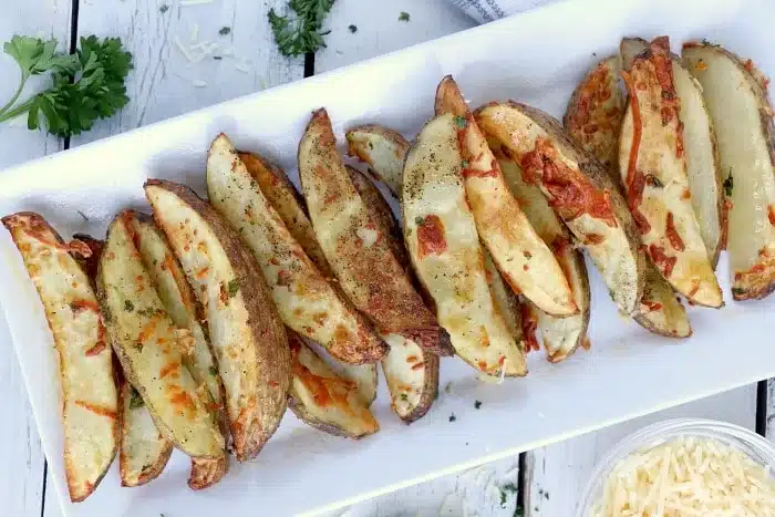 A rectangular white plate holds crispy air fryer potato wedges sprinkled with herbs and cheese. The plate rests on a white wooden surface, accompanied by a small bowl of grated cheese and a sprig of parsley nearby.