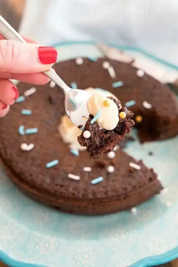 A hand with red nail polish holds a fork with a bite of instant pot chocolate cake topped with whipped cream and sprinkles. The cake, partially eaten, is on a decorative blue plate and is garnished with white and blue sprinkles.