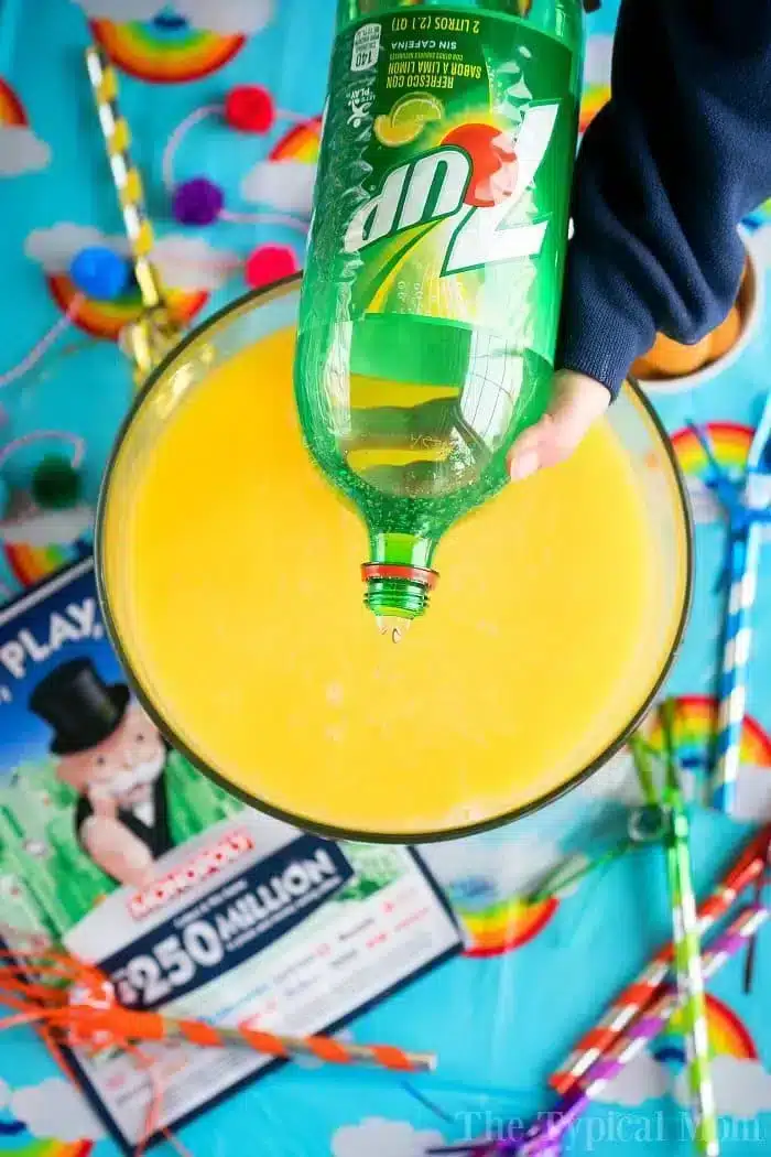 A hand pours 7UP into a bowl filled with a yellow liquid, crafting a festive party punch recipe. The table is adorned with a rainbow-themed tablecloth, colorful straws, and a Monopoly game board.