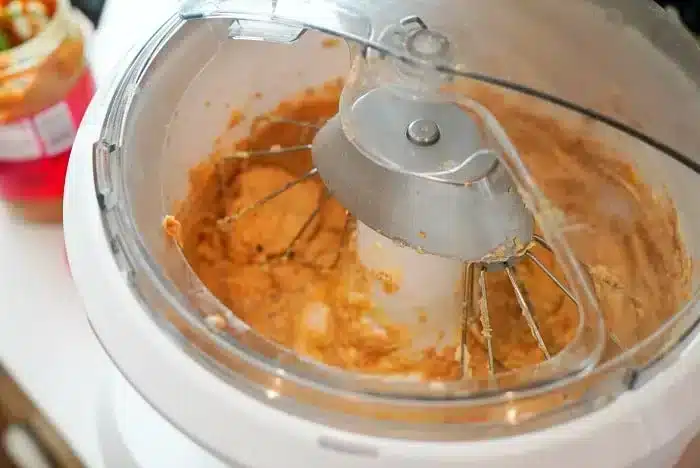 A close-up of a stand mixer with a whisk attachment mixing a thick, peanut butter-like substance. The surrounding area includes a partially visible jar with a red lid. Traces of the mixture cover the bowl and whisk attachment, hinting at the start of a delicious peanut butter oatmeal cookie recipe.