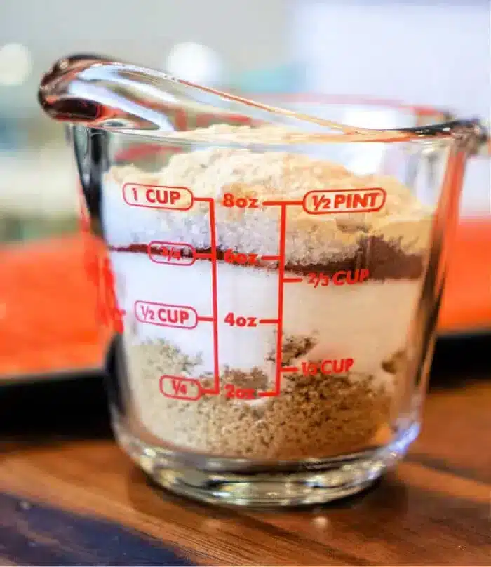 A clear measuring cup filled with layered ingredients, including brown sugar, flour, and a touch of salmon dry rub. The cup features red measurement markings for cups, ounces, and pints. It rests on a wooden surface.