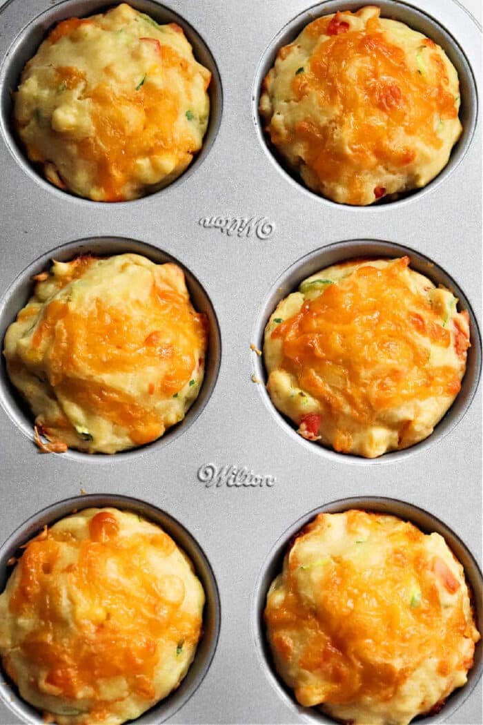 A close-up view of a muffin tin containing six savory veggie muffins. The muffins are topped with melted cheddar cheese, and their filling appears to include diced vegetables. The metallic muffin tin is labeled "Wilton.