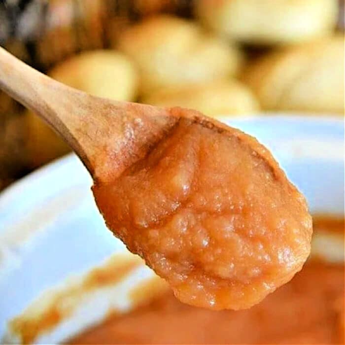 A wooden spoon holding a spoonful of thick, reddish-brown slow cooker apple butter above a white bowl filled with the same sauce. The background shows out-of-focus, round baked items.