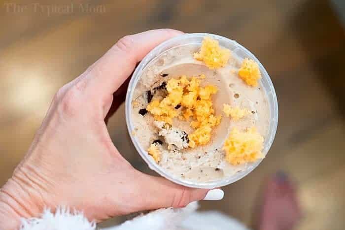 A person is holding a clear plastic cup filled with a creamy mixture, topped with crumbled orange 2 ingredient soda cake. The background is blurred, showing a wooden floor. The person's hand and part of a white fuzzy sleeve are visible.