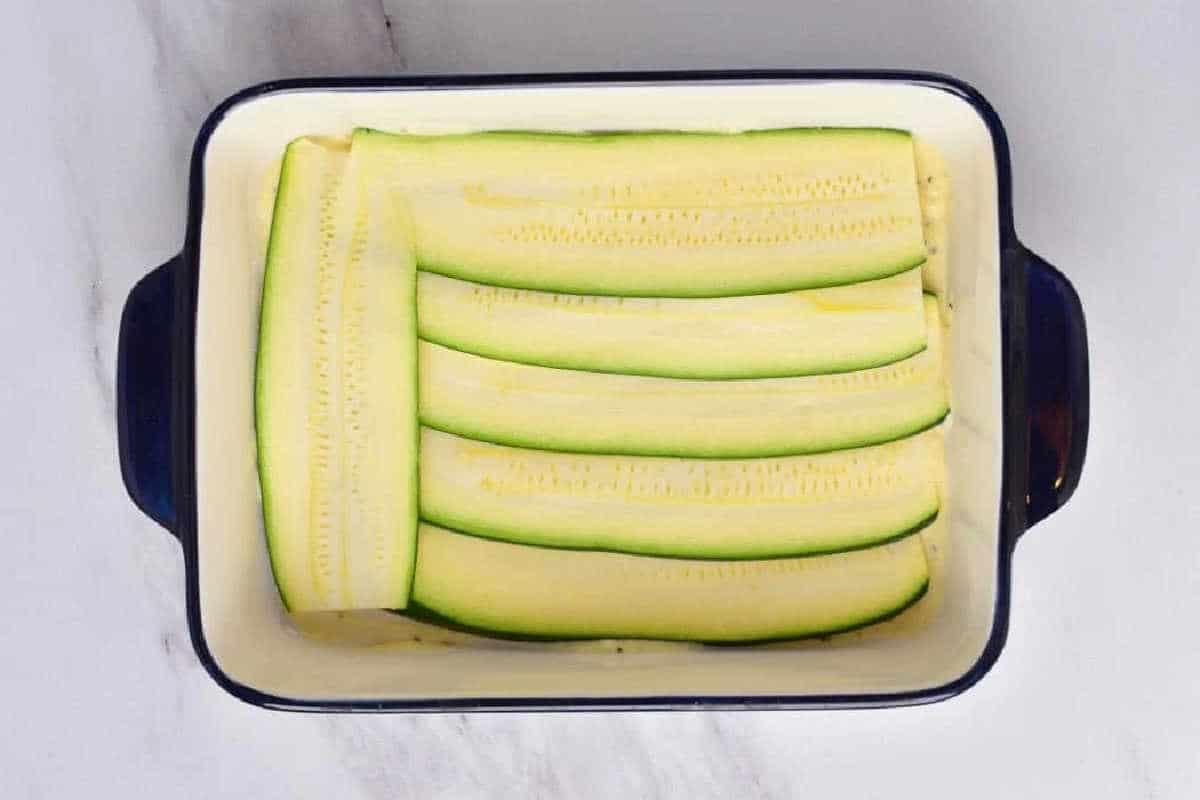 Rectangular dish with thin, raw zucchini slices neatly layered inside, ready to become a delicious vegetable lasagna with white sauce.
