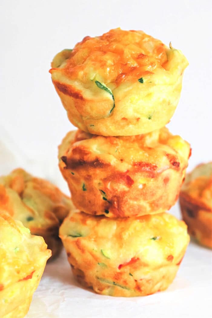 Three savory veggie muffins are stacked on top of each other against a plain white background. The muffins have a golden-brown top and are studded with green and orange vegetable bits. More muffins are partially visible at the sides.