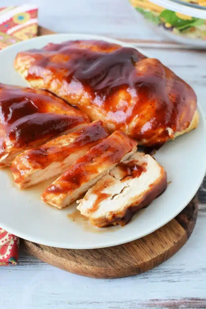 Sliced and whole air fryer BBQ-glazed chicken breast is served on a white plate. The plate rests on a wooden board, with part of a colorful napkin and bowl visible in the background.