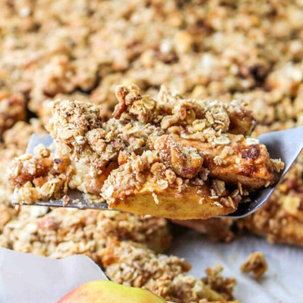 A spatula holds a freshly baked apple crumble piece, the perfect showcase for your apple slices recipe, with a whole apple in the foreground.