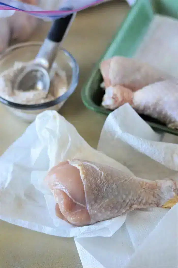 Raw chicken drumsticks are on white paper towels, ready for preparation. A small scoop rests in a bowl nearby containing a flour mixture. A green tray in the background holds additional chicken drumsticks, soon to become baked chicken legs.