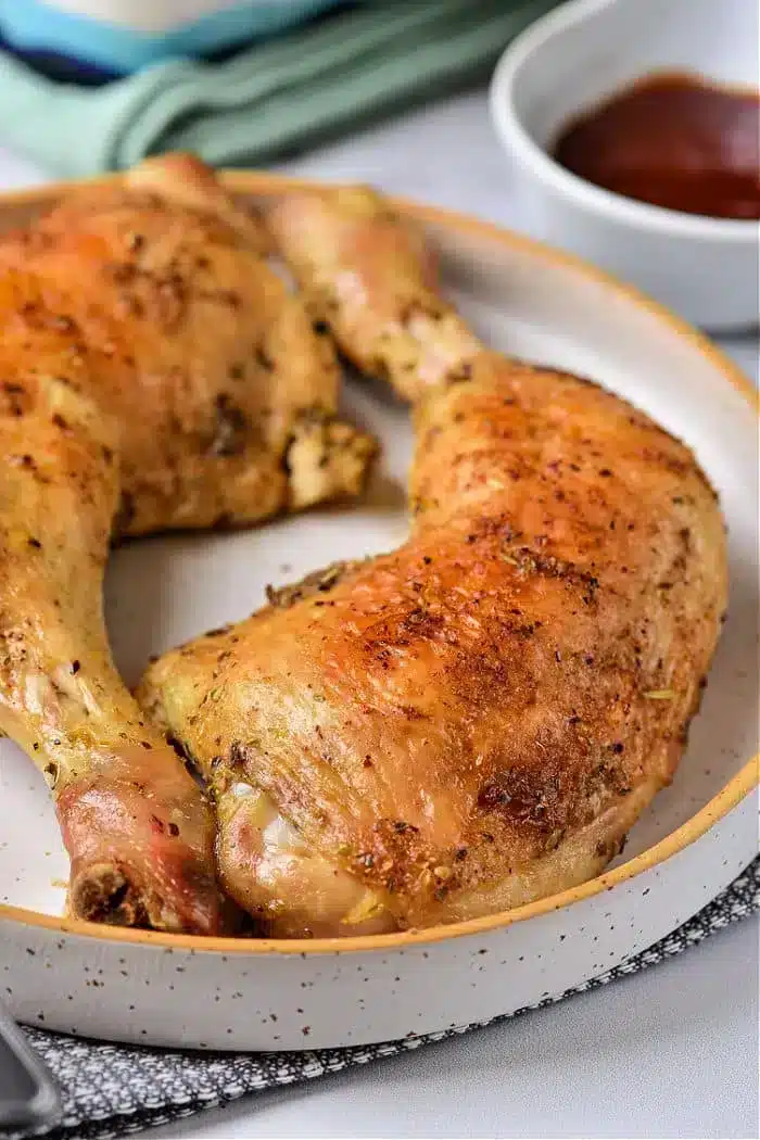 A plate containing two seasoned and cooked baked chicken legs. The chicken has a golden-brown, crispy skin. A bowl with a red dipping sauce is partially visible in the background. The plate is placed on a woven mat.