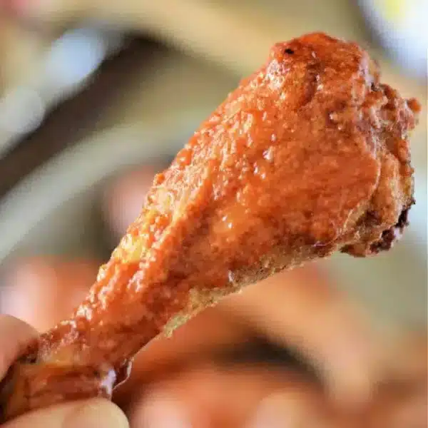 A close-up image of a hand holding a cooked chicken drumstick coated in a red sauce. The background appears to show more baked chicken legs on a plate, with blurred objects in the distance.