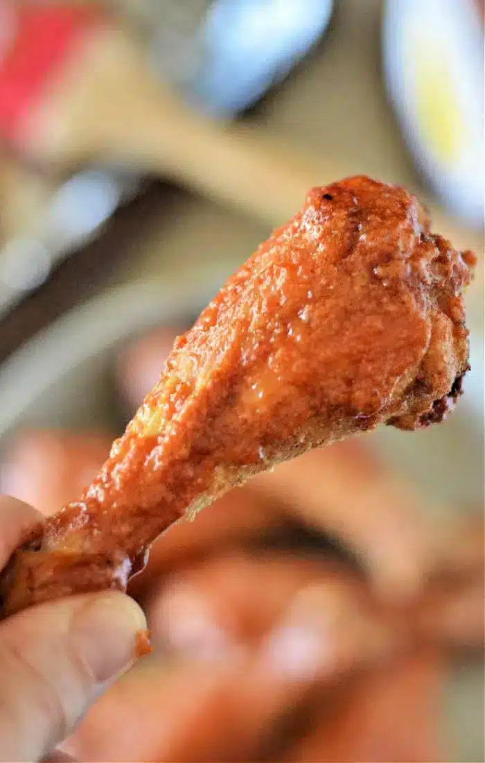 A close-up image of a hand holding a cooked chicken drumstick coated in a red sauce. The background appears to show more baked chicken legs on a plate, with blurred objects in the distance.