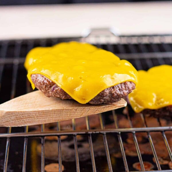 Baked hamburger patties with melted cheese sizzle on a grill, one being lifted with a wooden spatula.