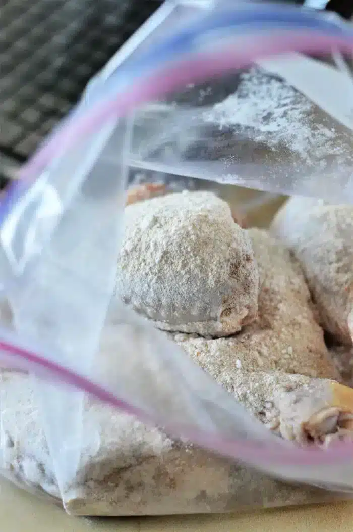 An open plastic bag containing raw, floured chicken pieces rests on a kitchen counter. The chicken pieces are coated with flour, ready for further preparation like turning them into delicious baked chicken legs. The background shows a partial view of a dark kitchen appliance.