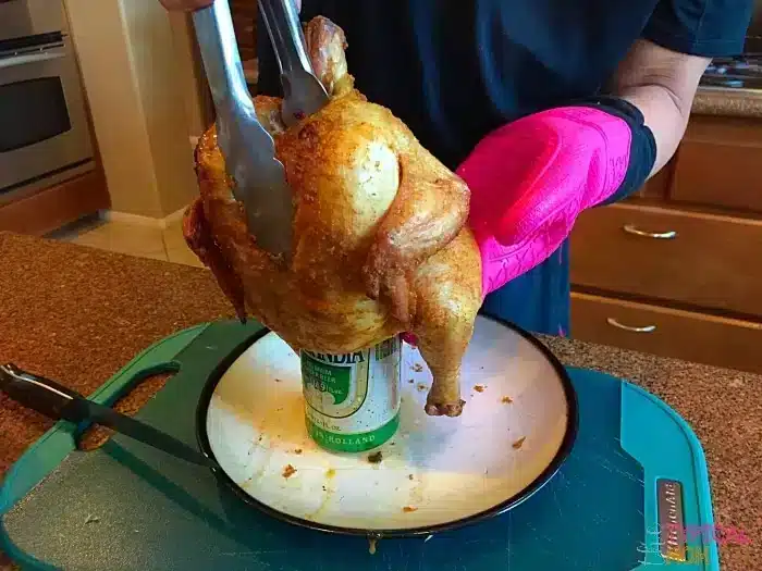 A person in a pink oven mitt uses tongs to lift a roasted beer can chicken from a charcoal grill. The bird sits proudly on a white plate atop a cutting board in the kitchen, with a knife ready for carving.