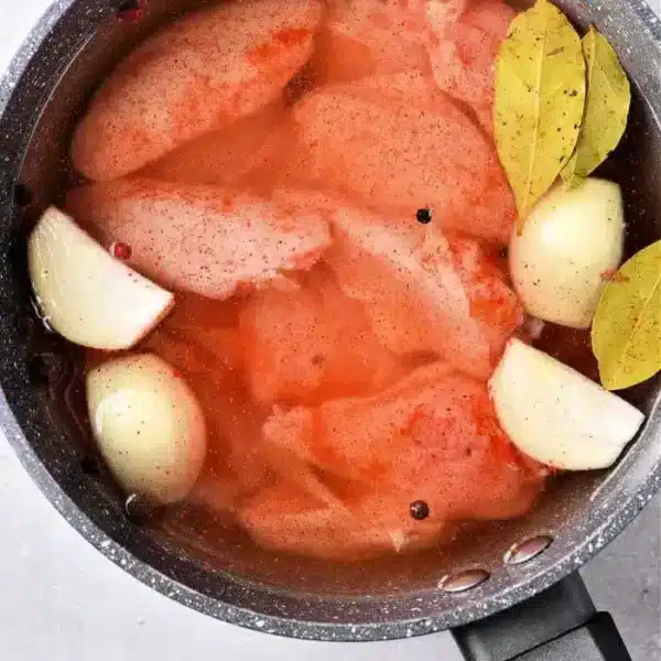 A pot containing raw chicken pieces, including boiled chicken wings, quartered onions, bay leaves, and whole peppercorns submerged in a reddish liquid, possibly broth or marinade, rests on a gray surface.