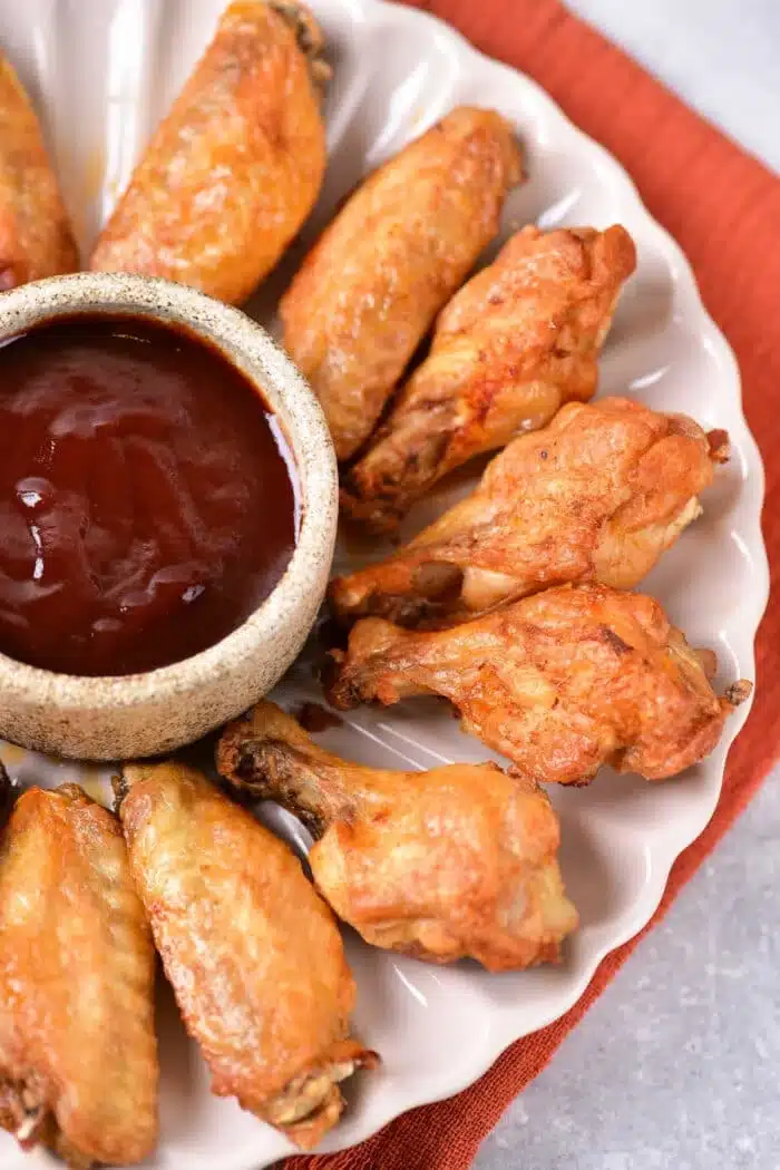 A plate of golden-brown, boiled chicken wings is arranged in a circular pattern around a small bowl filled with dark barbecue sauce. The plate rests elegantly on an orange cloth.