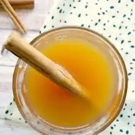 Top view of a crockpot hot apple cider in a glass, adorned with a cinnamon stick. The drink rests on a wooden surface beside a white cloth with green dots, while another cinnamon stick peeks onto the table.