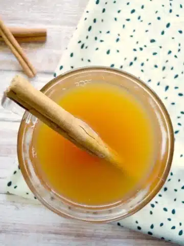 Top view of a crockpot hot apple cider in a glass, adorned with a cinnamon stick. The drink rests on a wooden surface beside a white cloth with green dots, while another cinnamon stick peeks onto the table.