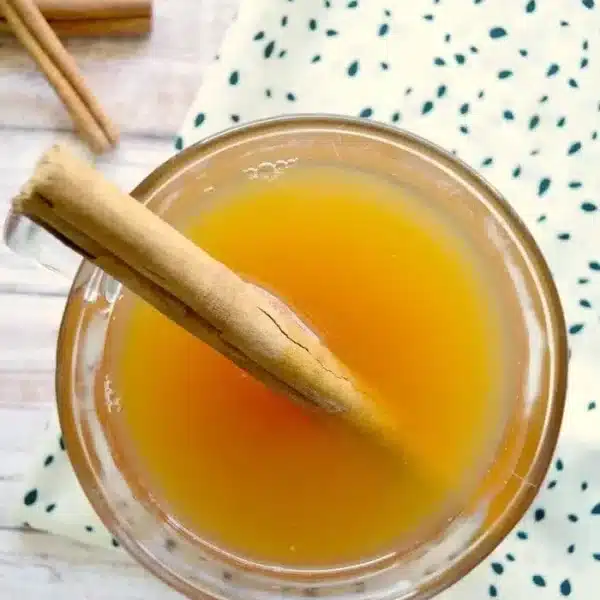 Top view of a crockpot hot apple cider in a glass, adorned with a cinnamon stick. The drink rests on a wooden surface beside a white cloth with green dots, while another cinnamon stick peeks onto the table.