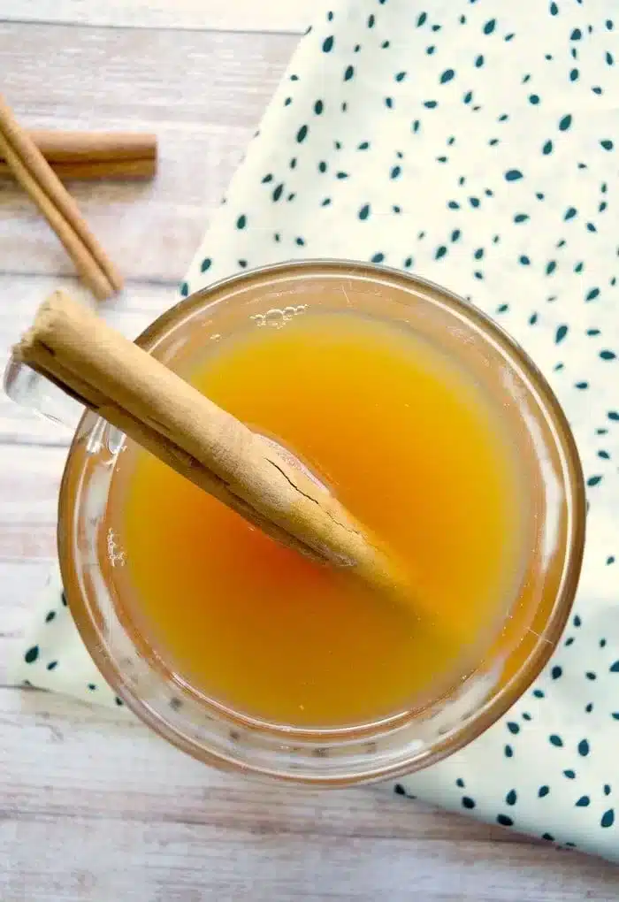 Top view of a crockpot hot apple cider in a glass, adorned with a cinnamon stick. The drink rests on a wooden surface beside a white cloth with green dots, while another cinnamon stick peeks onto the table.