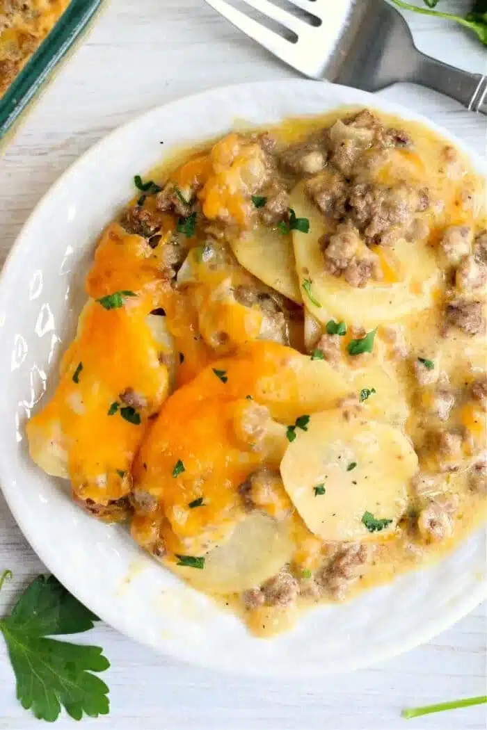 A plate of cheesy scalloped potatoes with ground beef, reminiscent of a classic Hamburger HotDish, garnished with chopped parsley. A fork and a sprig of parsley are visible nearby on the table.