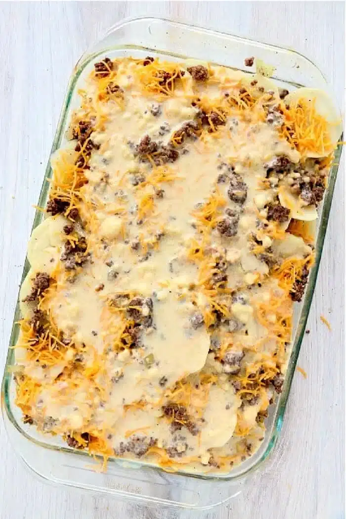 A glass baking dish filled with a delicious Hamburger HotDish showcases its layered casserole. Visible ingredients include sliced potatoes, browned ground beef, melted cheese, and a creamy sauce. The dish rests on a light-colored wooden surface, inviting you to savor its flavors.