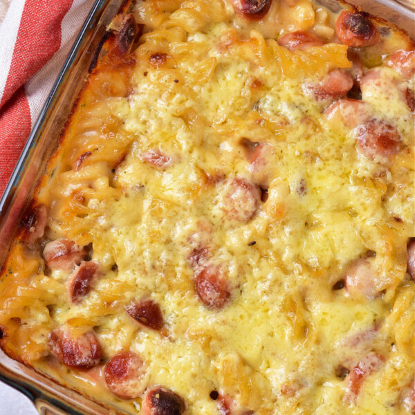 A baked dish in a glass pan, this hot dog casserole boasts a golden crust with visible slices of sausages and pasta, possibly macaroni. A red and white cloth is partially visible on the side.