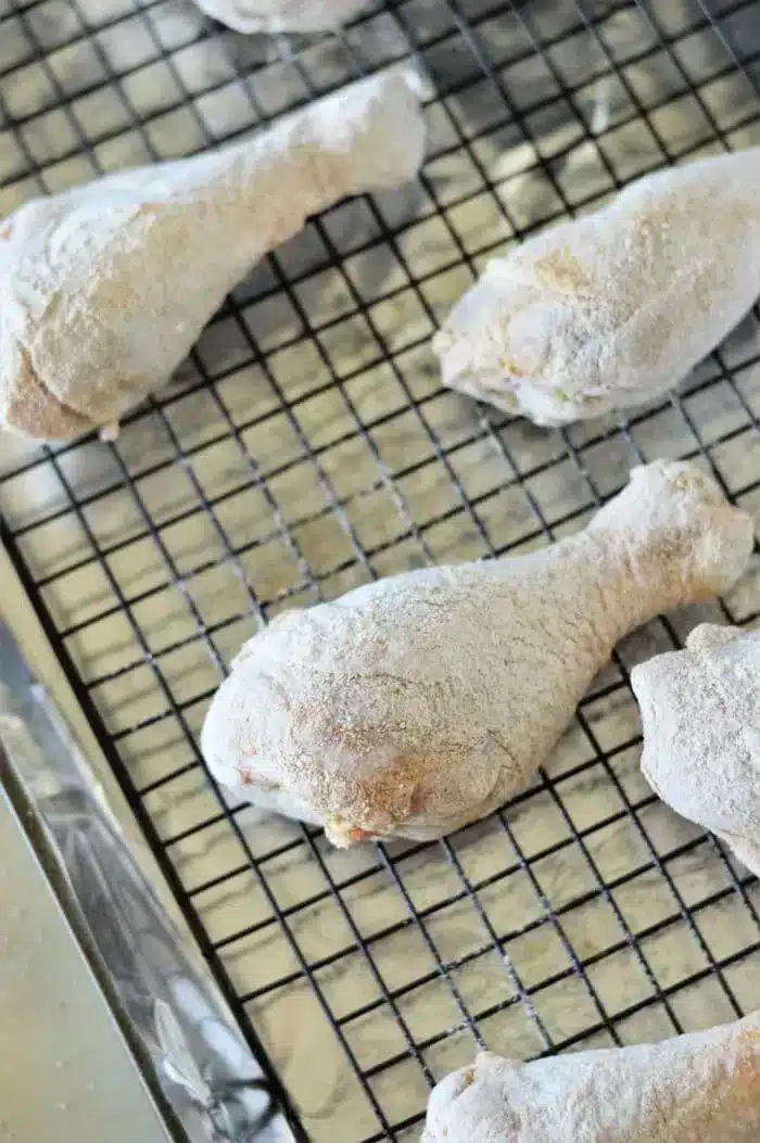 Baked chicken legs are coated in flour and placed on a wire rack for drying. The rack sits on a baking sheet lined with foil, suggesting the drumsticks are being prepared for frying or baking.