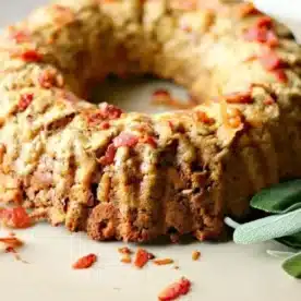 A savory bundt cake, shaped perfectly by the bundt pan, boasts a golden-brown crust and is topped with crispy bacon bits. Fresh sage leaves are artfully placed beside it on a beige surface, enhancing its rustic charm.