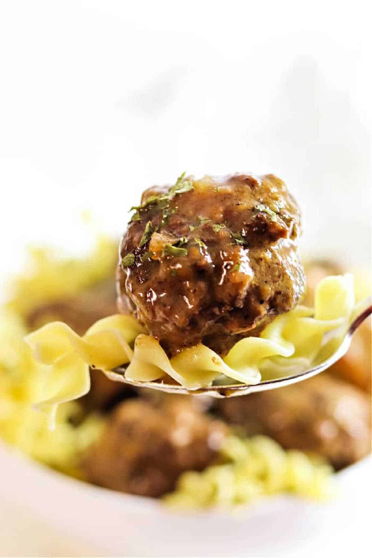 Close-up of a fork with a meatball and noodles, garnished with herbs, evokes the comforting flavors of Crockpot Meatballs and Gravy.