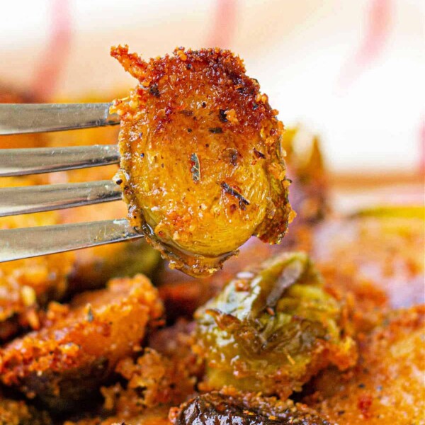 Close-up of a fork holding a crispy, Parmesan-crusted Brussels sprout slice with more on a white and blue-rimmed plate.