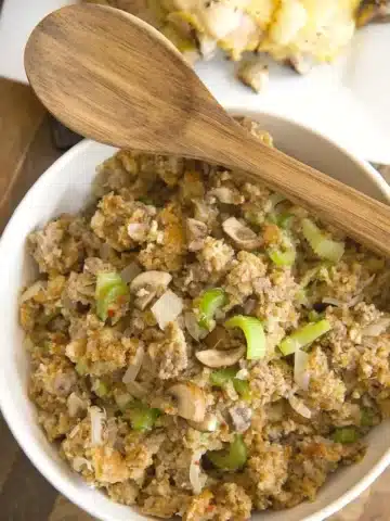 A bowl of stuffing featuring crumbled bread, sliced mushrooms, and chopped celery, with a wooden spoon resting on top. The delightful aroma hints at the Pepperidge Farm Sausage Stuffing Recipe. A dish of chicken is partially visible in the background.