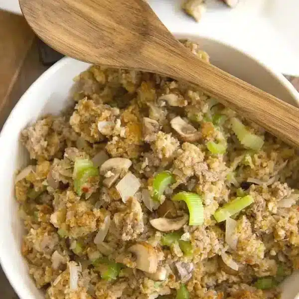 A bowl of stuffing featuring crumbled bread, sliced mushrooms, and chopped celery, with a wooden spoon resting on top. The delightful aroma hints at the Pepperidge Farm Sausage Stuffing Recipe. A dish of chicken is partially visible in the background.