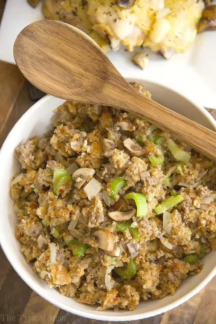 A bowl of stuffing featuring crumbled bread, sliced mushrooms, and chopped celery, with a wooden spoon resting on top. The delightful aroma hints at the Pepperidge Farm Sausage Stuffing Recipe. A dish of chicken is partially visible in the background.