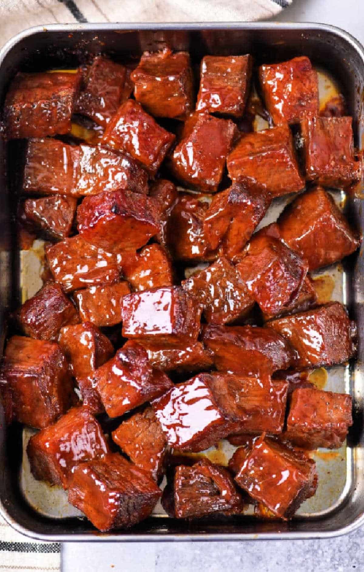 Close-up of glazed, cooked chuck roast burnt ends arranged in a metal baking tray.