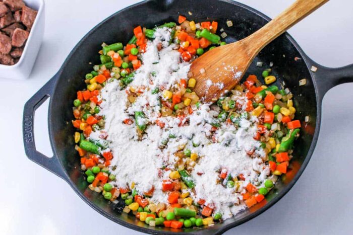 A skillet of mixed vegetables with flour being stirred by a wooden spoon, perfect for starting a delicious Leftover Steak Pot Pie.