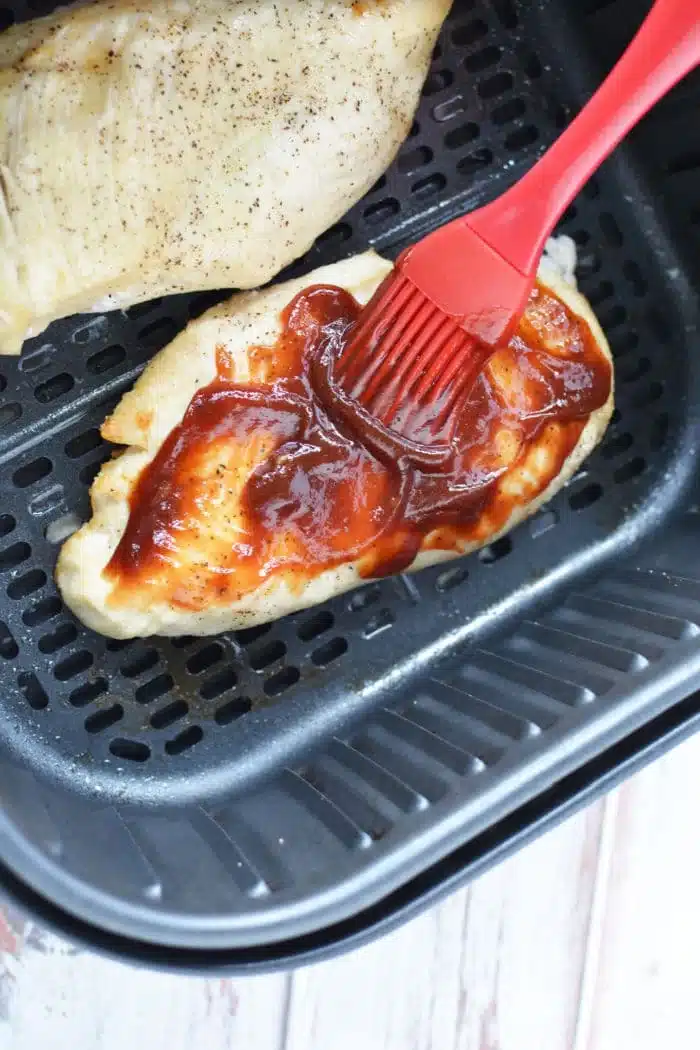 A piece of air fryer BBQ chicken is being brushed with barbecue sauce using a red silicone brush inside the basket. Another piece of seasoned chicken sits tantalizingly in the background, ready to be savored.