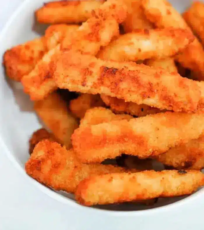 A white bowl filled with multiple crispy, golden-brown air fryer fish sticks, arranged in a slightly haphazard manner. The breaded fish sticks appear to be freshly cooked, with a crunchy texture visible. The background is plain and white.