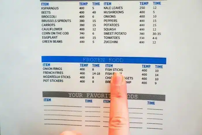 A person is pointing at a section of a cooking chart titled "FROZEN FOOD." The chart lists cooking times and temperatures for various vegetables and frozen foods, including air fryer fish sticks and chicken nuggets. 