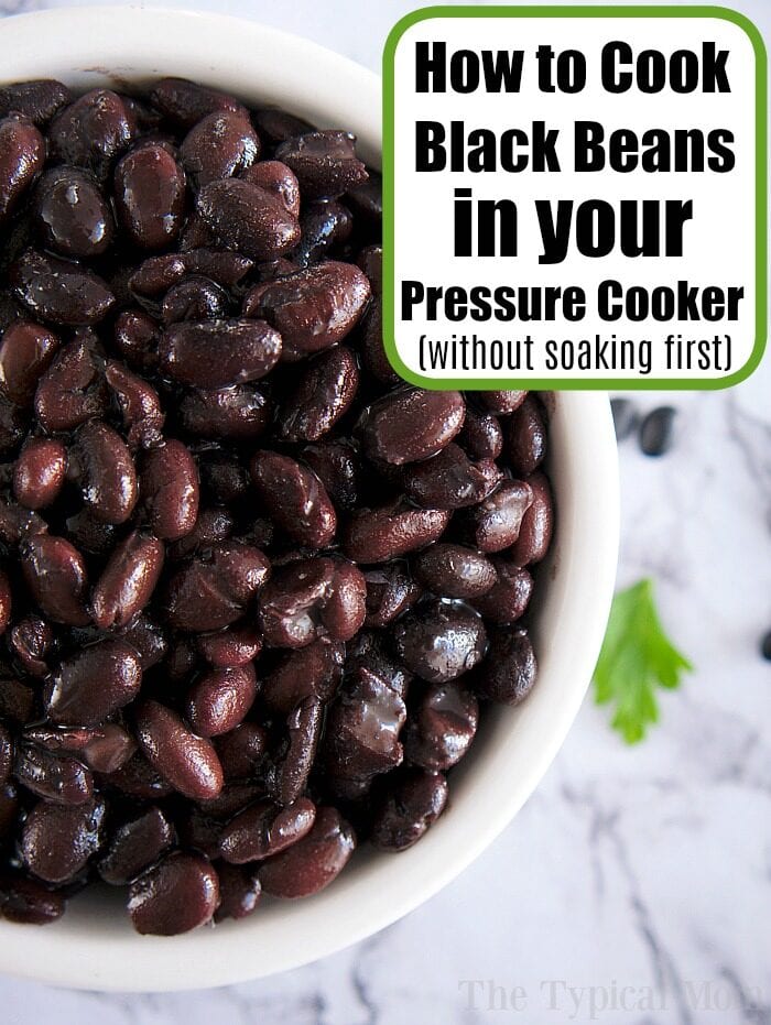 A bowl of cooked black beans rests on a marble surface, perfect for batch cooking. A text overlay reads, "How to Cook Black Beans in your Pressure Cooker (without soaking first)." A sprig of cilantro is visible beside the bowl.