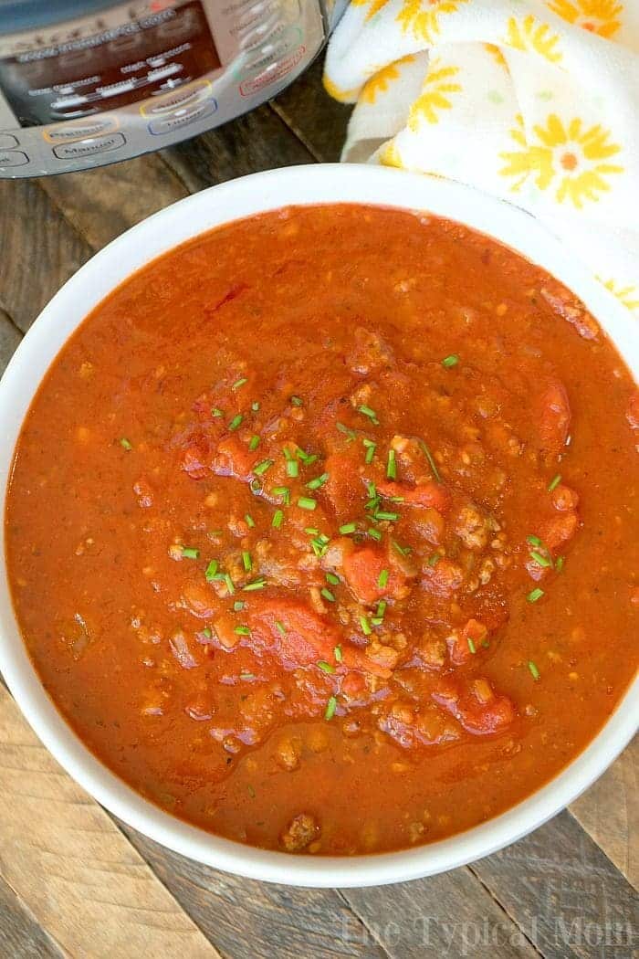 A white bowl filled with chunky tomato and meat stew, perfect for batch cooking, is garnished with chopped herbs and placed on a wooden table. A yellow and white patterned cloth and a pressure cooker are partially visible in the background.