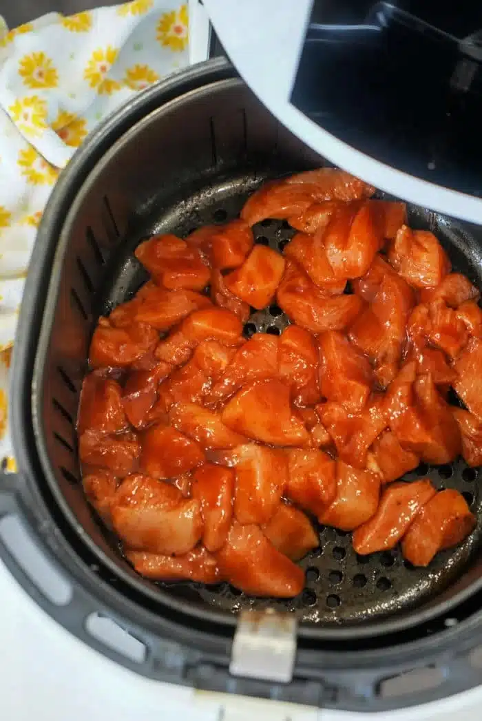 Chunks of marinated chicken are placed inside an open air fryer basket, sizzling into succulent air fryer BBQ chicken. A white kitchen towel with yellow floral patterns is partially visible on the side.