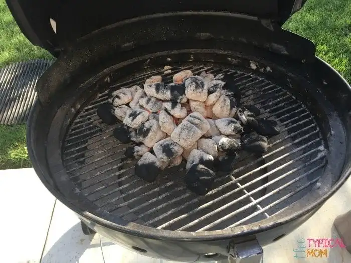 A round charcoal grill with its lid open reveals a pile of partially burned charcoal briquettes, perfect for preparing beer can chicken. The backdrop features some grass and patio tiles, setting the scene for a relaxed backyard barbecue.