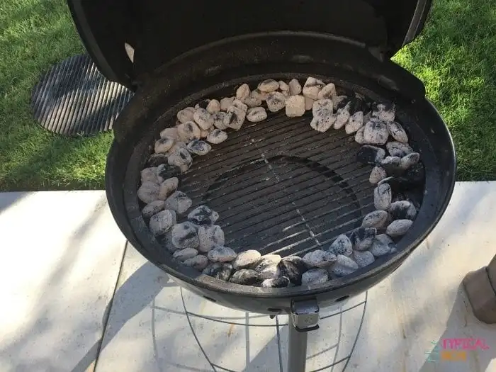 A round charcoal grill, ready for a classic beer can chicken, features unlit coals arranged in a circle along the outer edge. The center remains empty for culinary magic. Positioned on concrete, it's surrounded by a touch of greenery in the background.